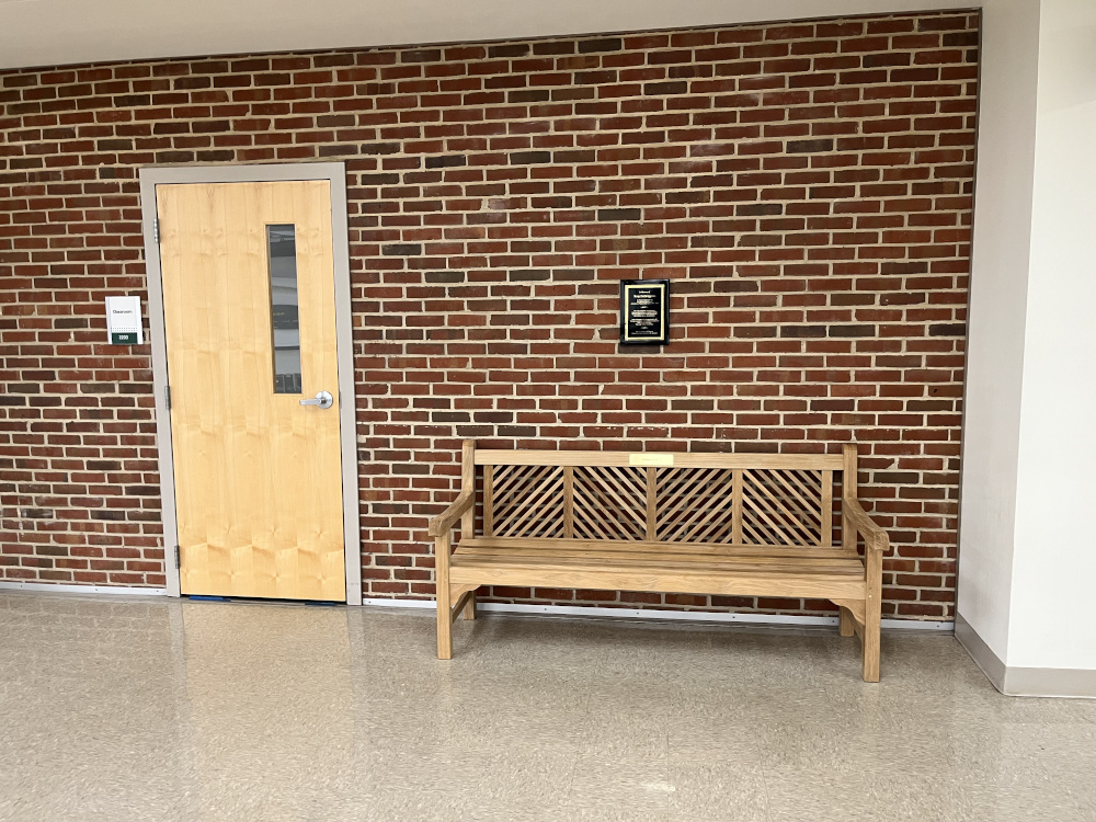 A photo of the bench by the classroom door. A plaque above the bench reads: In Memory of Tony DeGregorio. BS Physical Education '84, MS Physical Education '87, Instructor of Physical Education, 1990 - 2019. Mr. D was passionate about preparing future health and physical education teachers. He found great joy in nurturing relationships with students, alumni, and colleagues. Some of his memorable sayings loved by all: 'Set high expectations. Your students will rise to it.' 'Show me the good stuff.' 'Fun is the vehicle for learning.' Mr. D's Legacy will live on through the many educators he inspired.