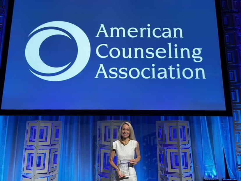 Stephanie Dailey holding the award on stage standing under a large logo and text saying American Counseling Association