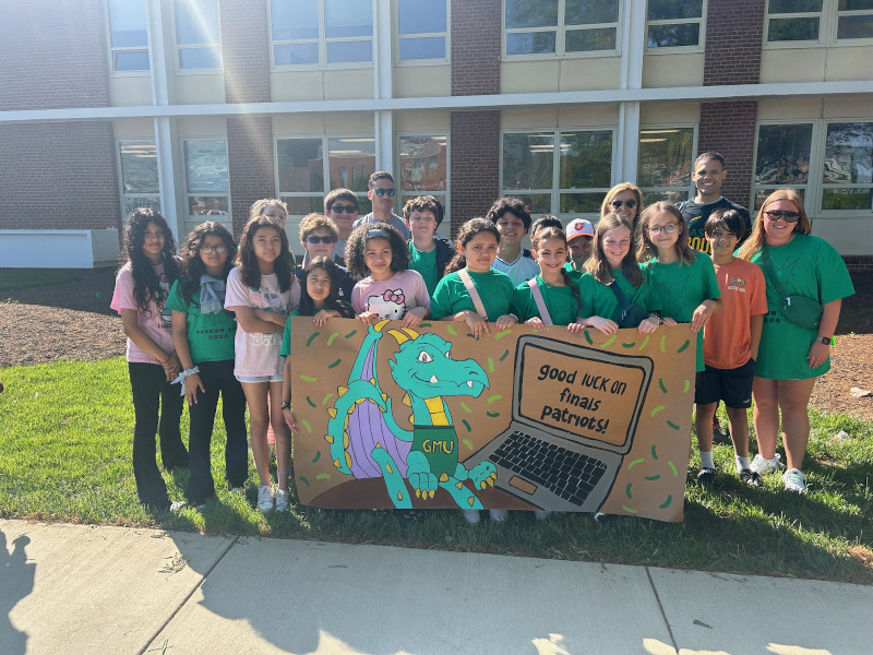 Students from Daniels Run Elementary School pose with a poster they painted featuring a dragon in a GMU shirt at a laptop with the message 'Good luck on finals patriots!'